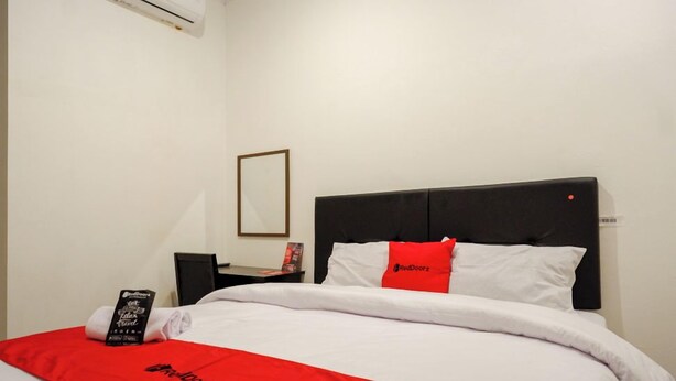 A neatly made bed with white and red bedding, a black headboard, and a small bedside table with a lamp and a book. - Reddoorz @ Jalan Wonosari Km.9 (Yogyakarta)