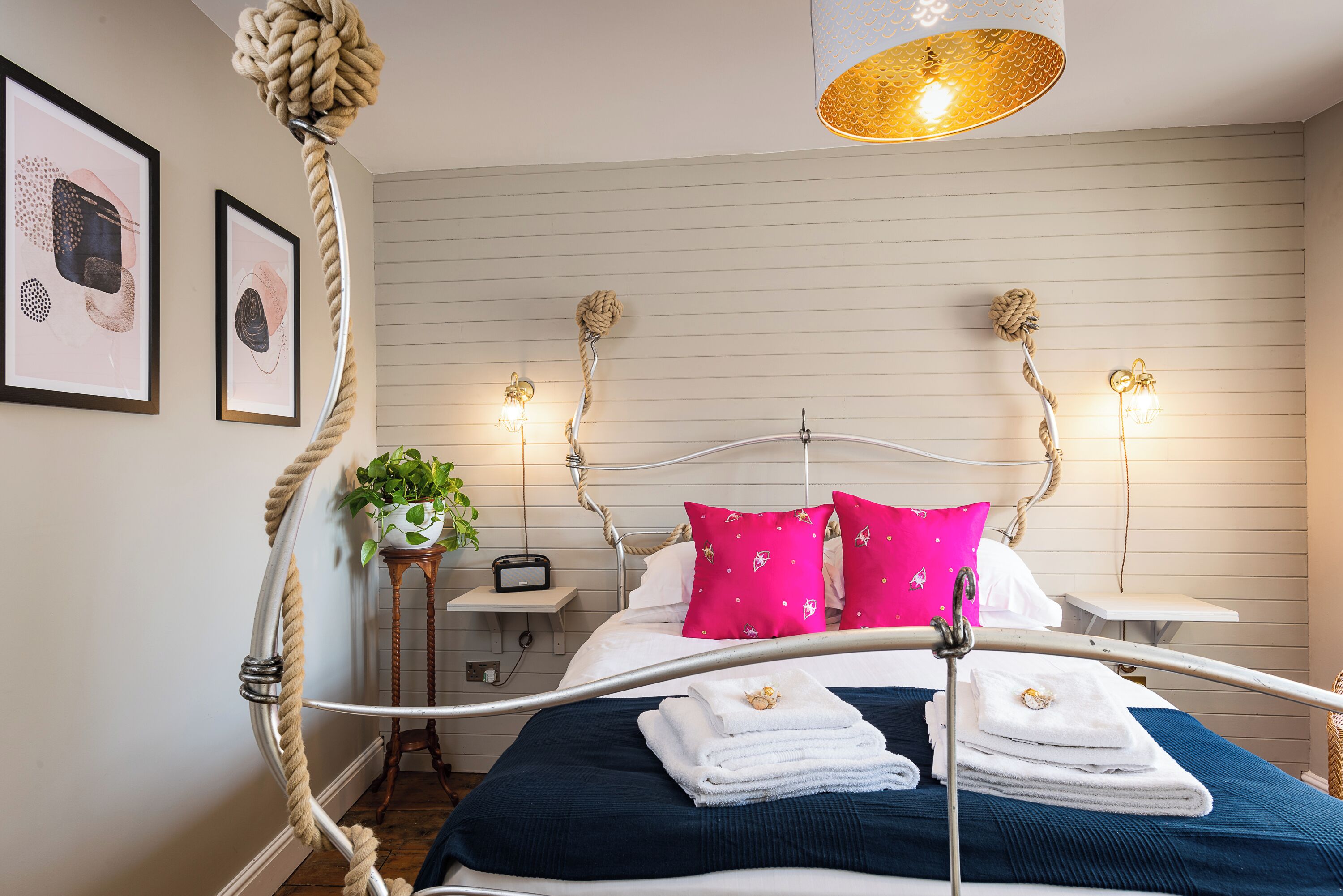 A neatly made bed with white and pink pillows, a blue bedspread, and a metal headboard. There are two bedside tables with lamps and a small plant on one.