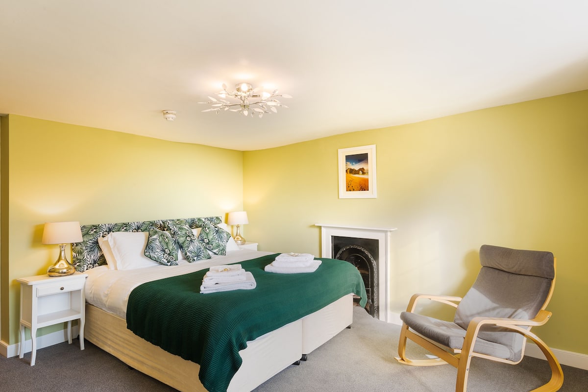A bedroom with a bed, bedside lamps, a green throw blanket, a chair, and a fireplace.