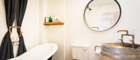 A bathroom with a freestanding bathtub, a toilet with a wooden seat, a round mirror, and a wooden barrel sink.