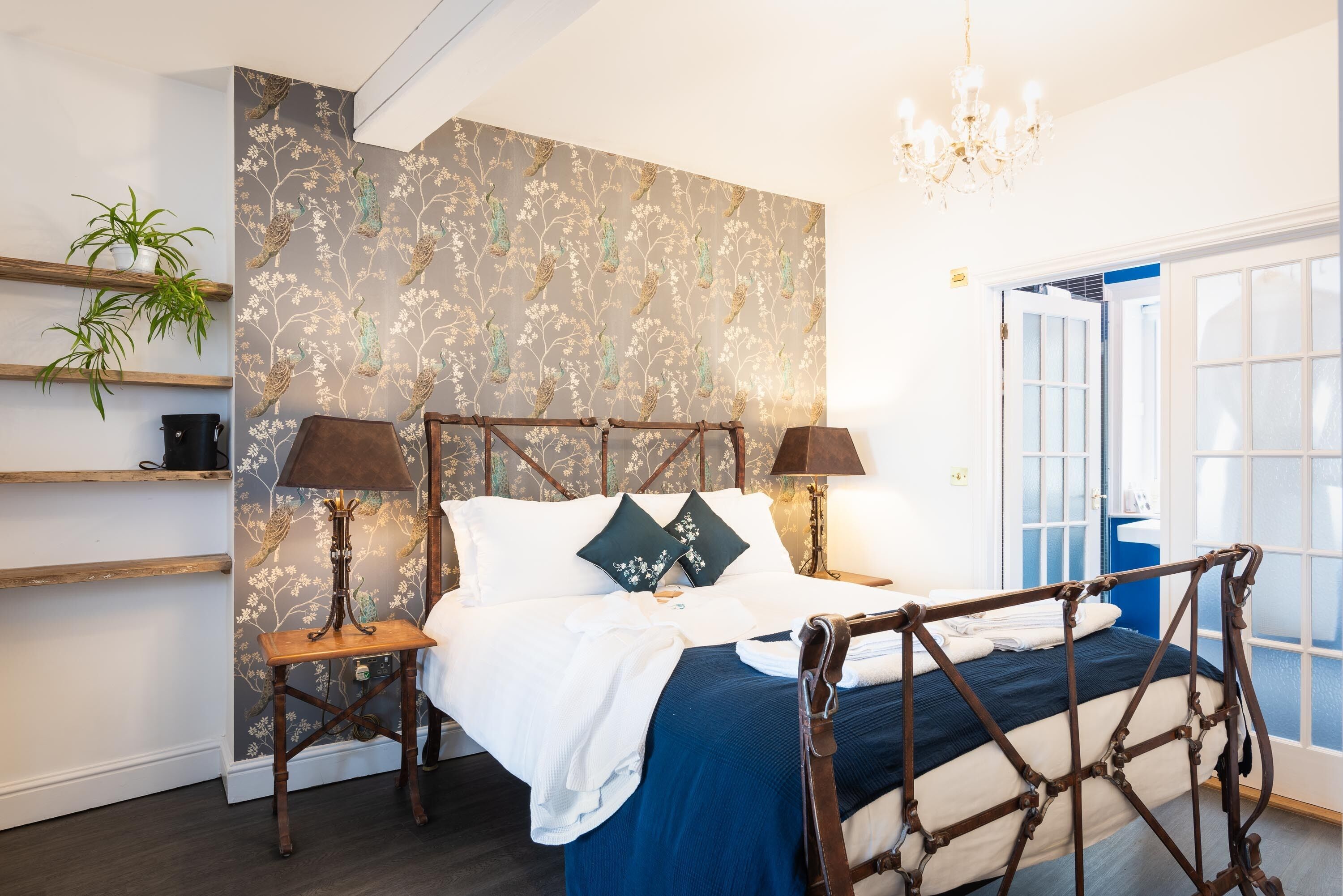 A bedroom with a bed, two bedside tables, a chandelier, a plant, and a window with a view.