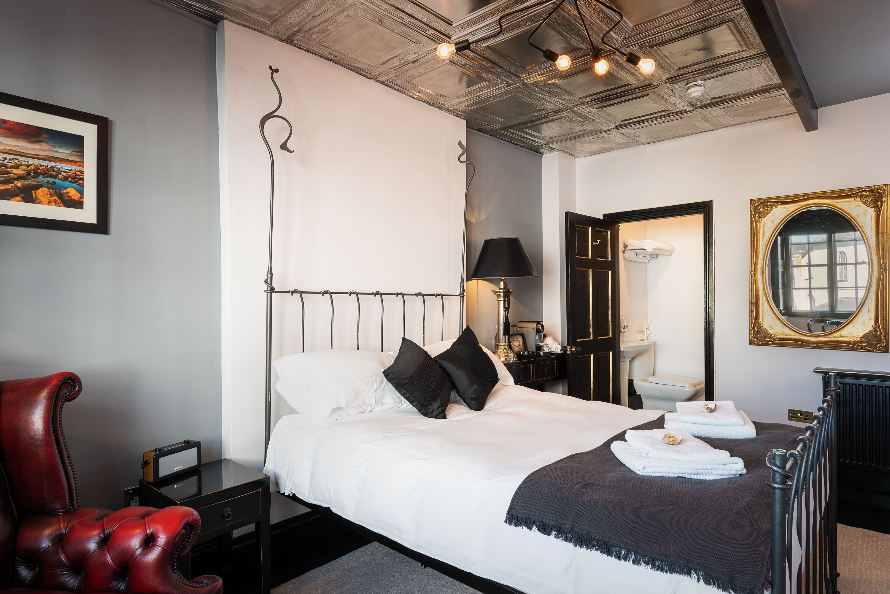 A neatly made bed with white linens and black pillows, a red leather chair, a small table with a lamp, and a framed picture on the wall.