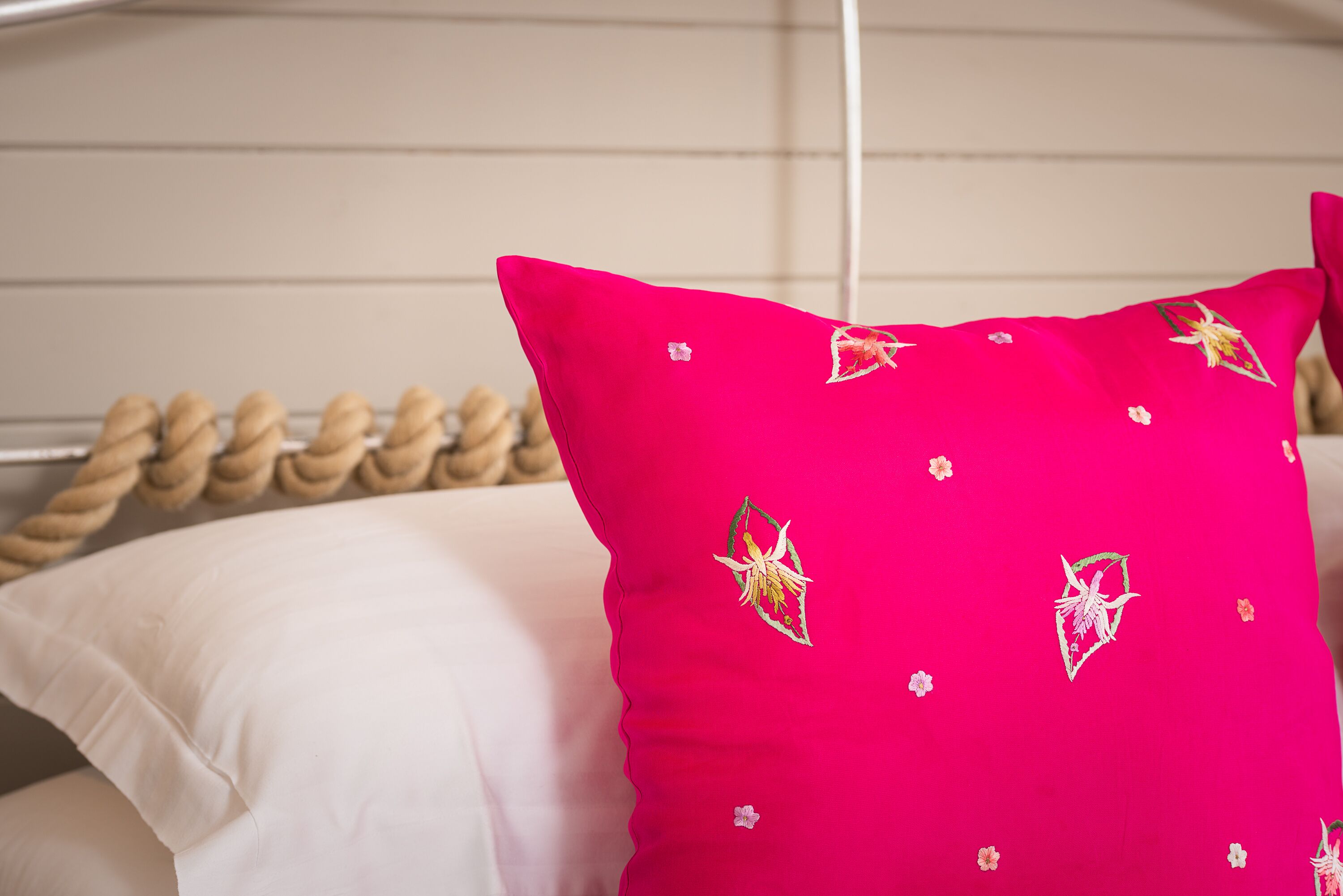 A bed with a white pillow and a bright pink pillow with floral embroidery.