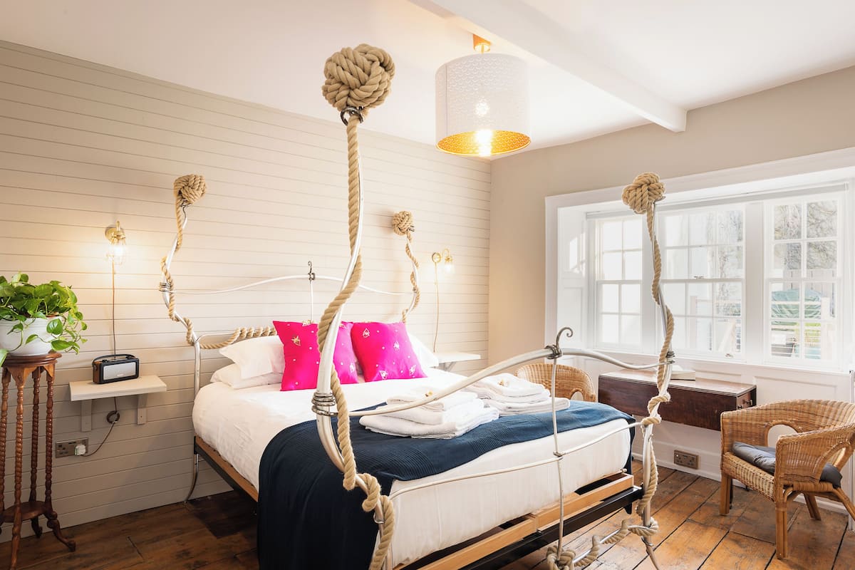 A four-poster bed with rope decorations, a bedside table with a radio, a wicker chair, and a large window with a view.