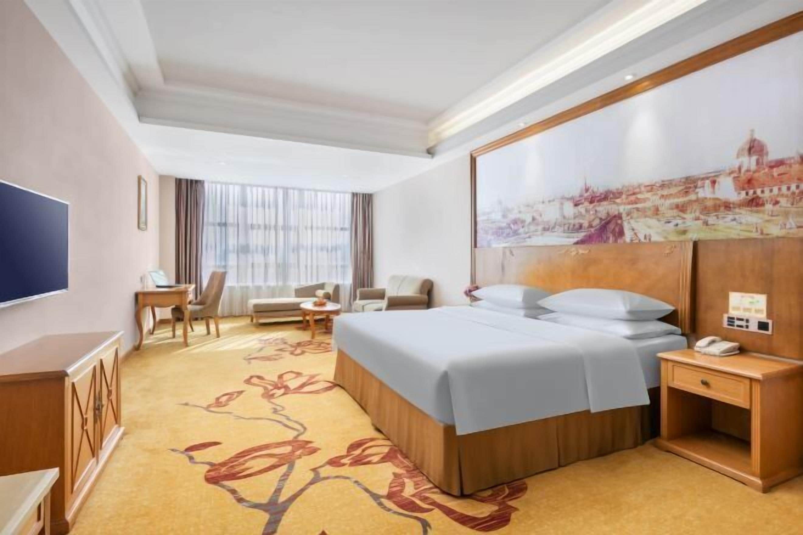 A hotel room with a large bed, a desk, a chair, a television, and a painting on the wall.