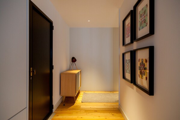 Un pasillo con suelo de madera, una puerta oscura, un pequeño armario de madera con una lámpara y cuadros en la pared.
