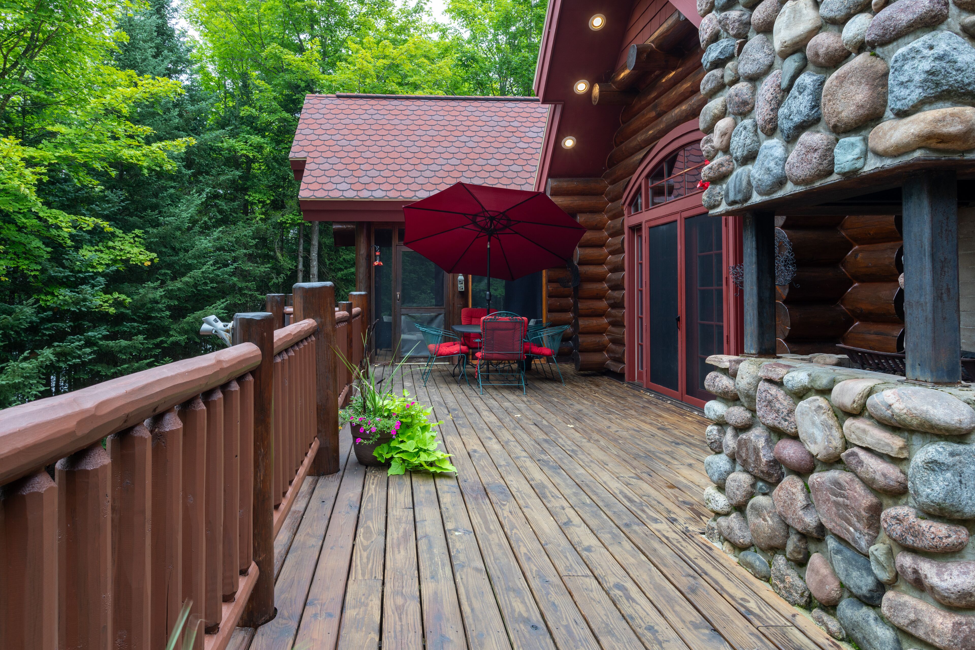 Una terraza de madera con un muro de piedra, un paraguas rojo y una mesa con sillas.