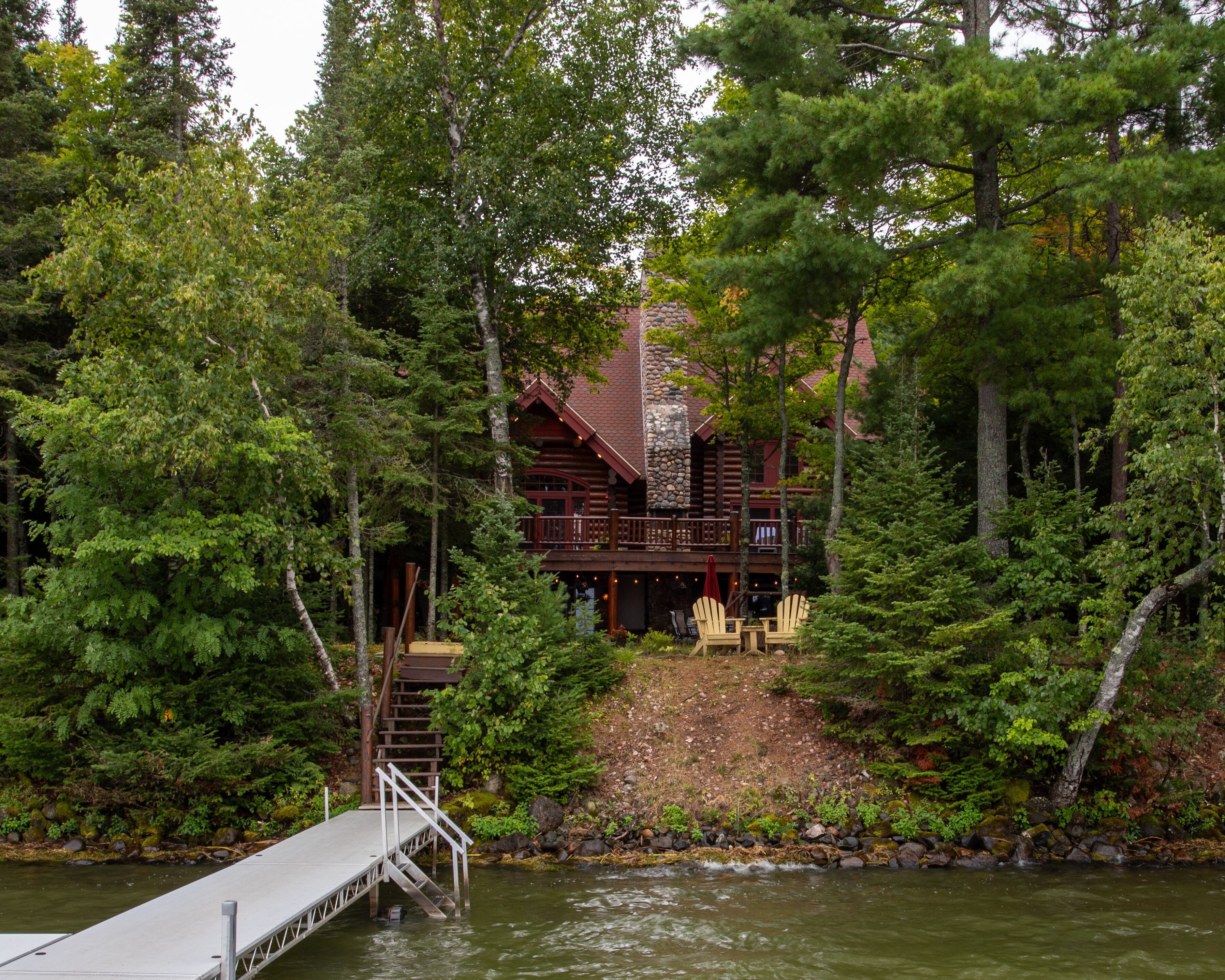 A log cabin with a stone fireplace, surrounded by dense forest and a dock leading to a small pier.