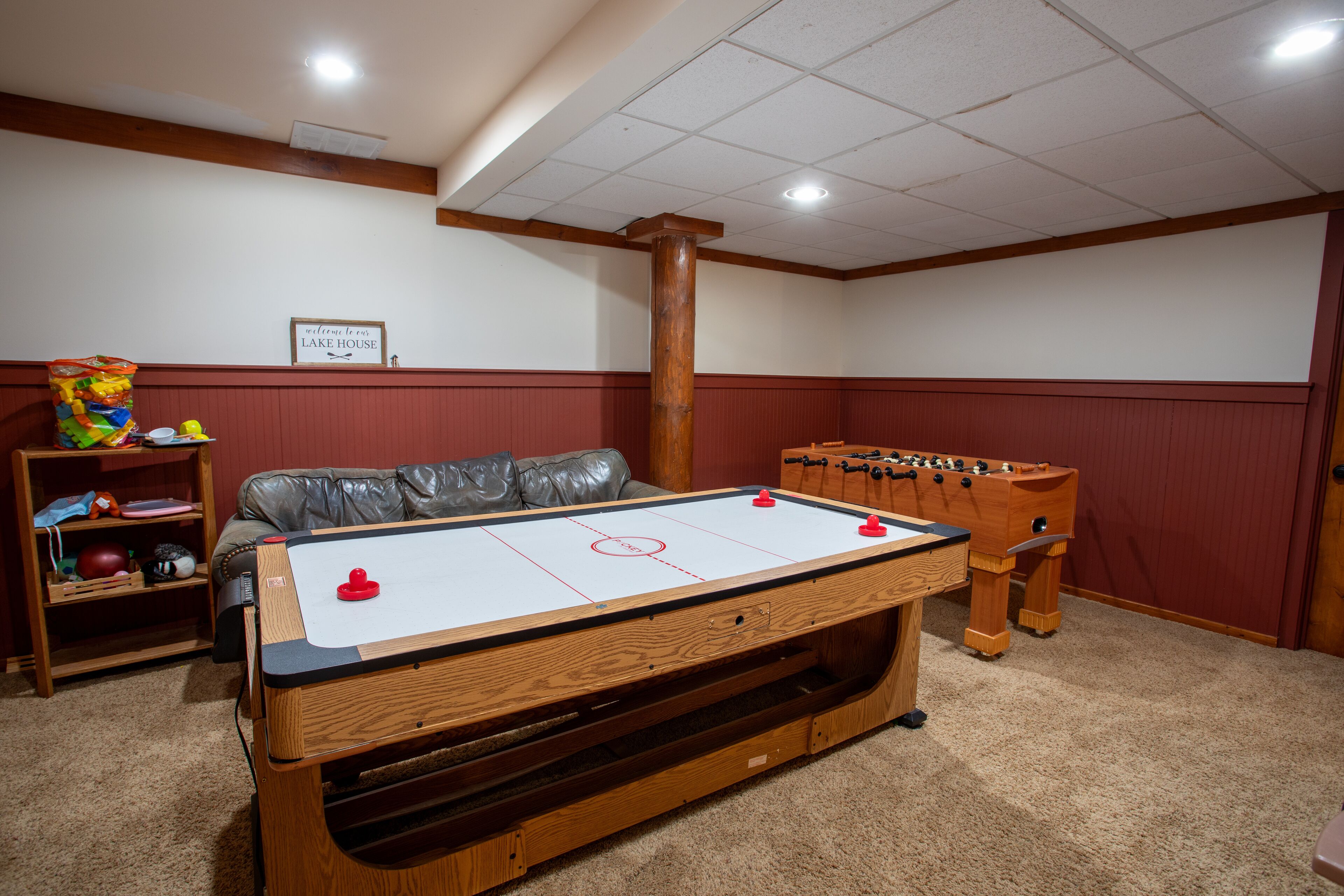 Una mesa de billar junto a una mesa de fútbol de mesa, un sofá de cuero y una estantería en una sala con paneles de madera y un cartel en la pared.