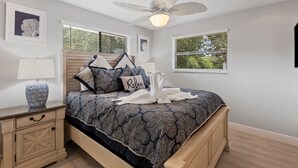 Ein Schlafzimmer mit einem Bett, einem Nachttisch, einer Lampe, einem Deckenventilator und zwei Fenstern mit Jalousien.
