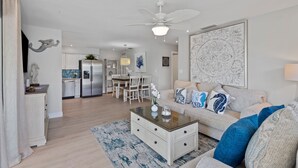 Ein modernes Wohnzimmer mit einer Couch, einem Couchtisch und dekorativen Kissen. Es gibt eine Essecke mit Tisch und Stühlen sowie eine Küche mit Geräten.
