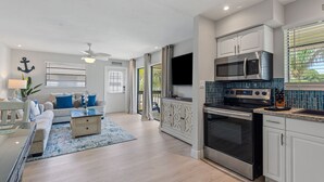 Eine moderne Küche mit Edelstahlgeräten, ein Essbereich mit blauem Teppich und ein Wohnzimmer mit Sofa und Deckenventilator.