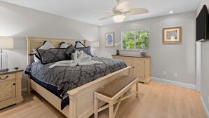 Une chambre à coucher comprenant un lit, un banc, une table de chevet, un ventilateur de plafond, une fenêtre donnant sur un paysage verdoyant et un tableau encadré accroché au mur.