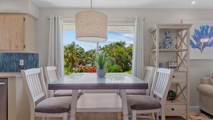 Un coin repas avec une table en verre, des chaises blanches, un luminaire suspendu et une vue sur des palmiers par la fenêtre.