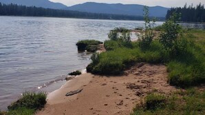 A lakeside view with a sandy beach, grassy areas, and mountains in the background.
