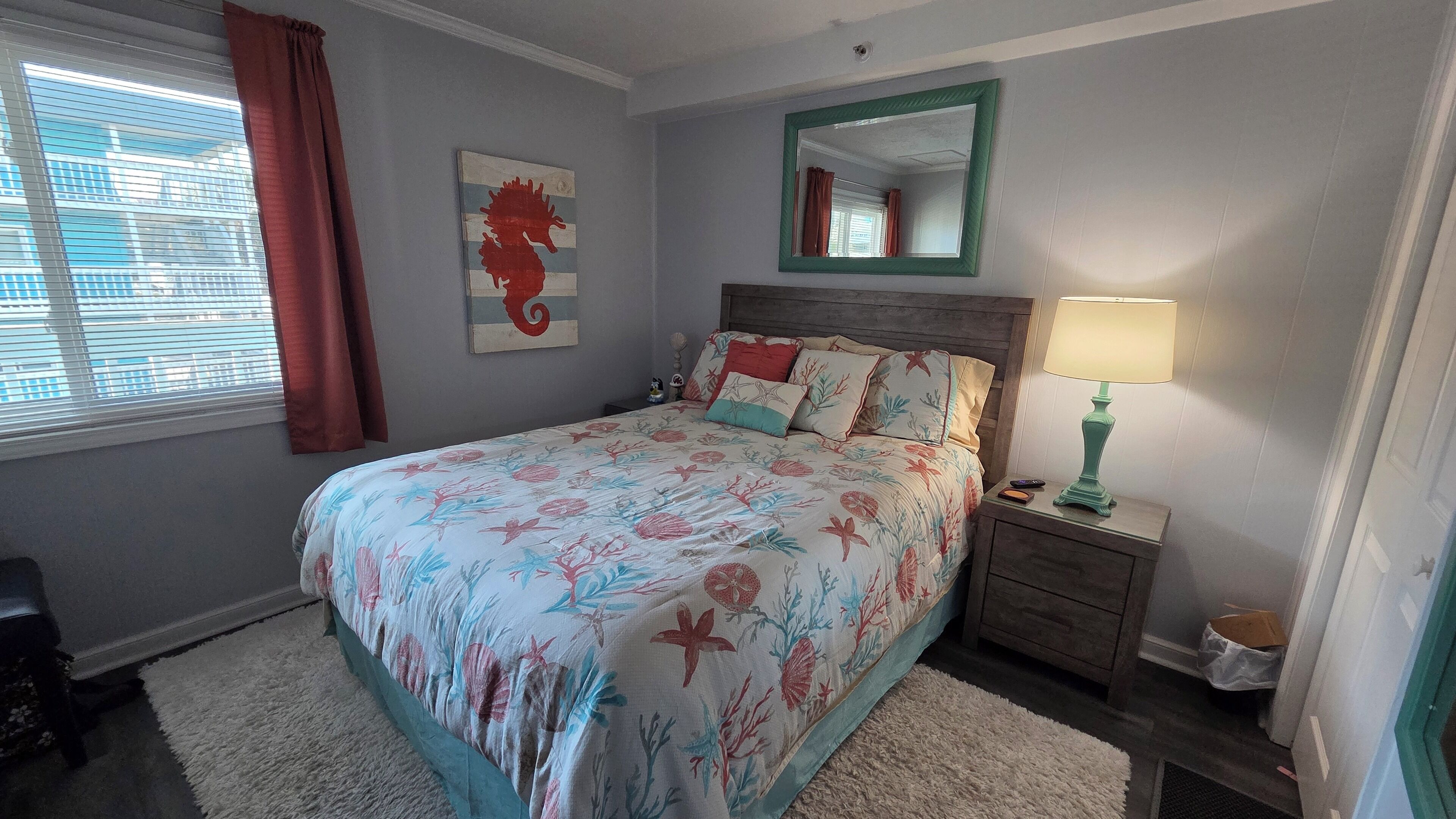 Un dormitorio con una cama, una mesita de noche, una lámpara, un espejo y una obra de arte con forma de caballito de mar en la pared.