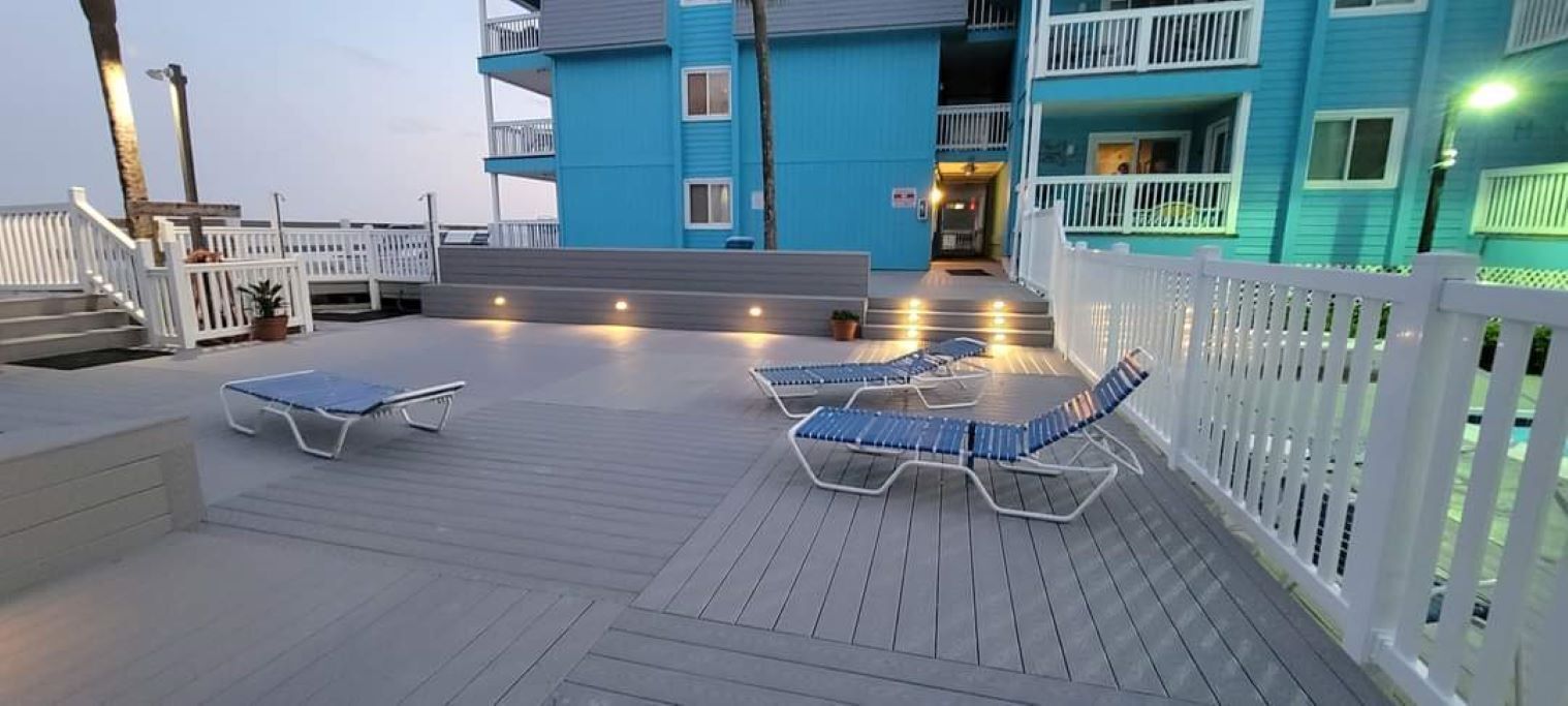 Une terrasse avec deux chaises longues et une petite table, une clôture blanche et, au fond, un bâtiment bleu doté de balcons.
