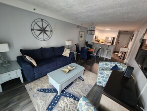 A living room with a dark blue sofa, a light blue coffee table, and patterned rugs. A kitchen is visible in the background with white cabinets and a dining area.
