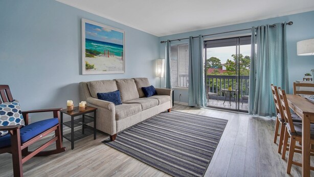 A living room with a sofa, a chair, a table, and a balcony with a view of the outdoors. - Unwind by the Beach - Savannah/Tybee (Tybee Island)