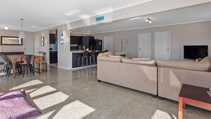 A modern living room with a large beige sofa, a coffee table, and a TV. There is a kitchen area in the background with a bar and stools. - Surf, Sand & Summer Vibes. (Waiotahe)