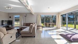 A spacious living room with a large TV, a sofa, and a dining area with a table and chairs. - Surf, Sand & Summer Vibes. (Waiotahe)