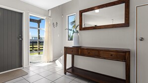 A hallway with a wooden console table, a large mirror, and a door leading to an outdoor area. - Surf, Sand & Summer Vibes. (Waiotahe)