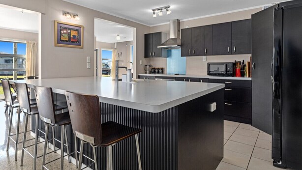 A modern kitchen with a central island, bar stools, and dark cabinetry. - Surf, Sand & Summer Vibes. (Waiotahe)