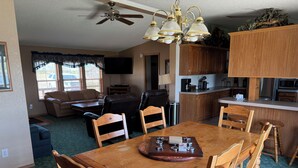 A dining area with a wooden table and chairs, a ceiling fan, and a kitchen in the background. - A Fisherman's Paradise (Harrold)