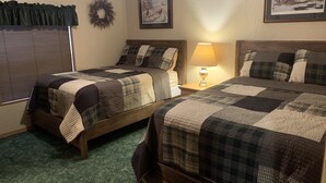 A bedroom with two beds, a nightstand with a lamp, a window with blinds, and a wall with a wreath and a framed picture. - A Fisherman's Paradise (Harrold)