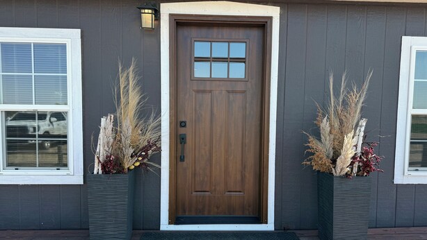 A house with a brown front door, two potted plants with dried plants, a doormat, and a window with blinds. - A Fisherman's Paradise (Harrold)