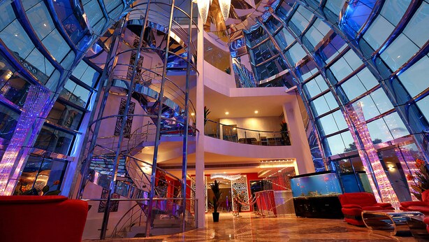 Un moderno vestíbulo de hotel con un atrio de cristal, varios pisos y una escalera central.