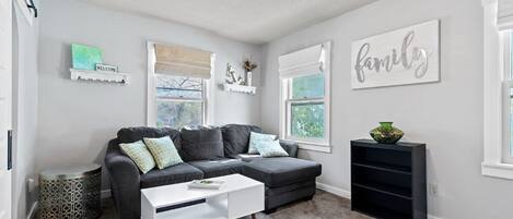 A living room with a grey sectional sofa, a white coffee table, and a black entertainment unit. There are two windows with white curtains, a ceiling fan, and a wall-mounted 'family' sign.