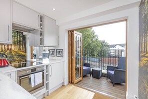 Una cocina moderna con horno integrado y un hervidor rojo, una puerta corrediza de vidrio que da a un área de estar exterior con muebles de mimbre.