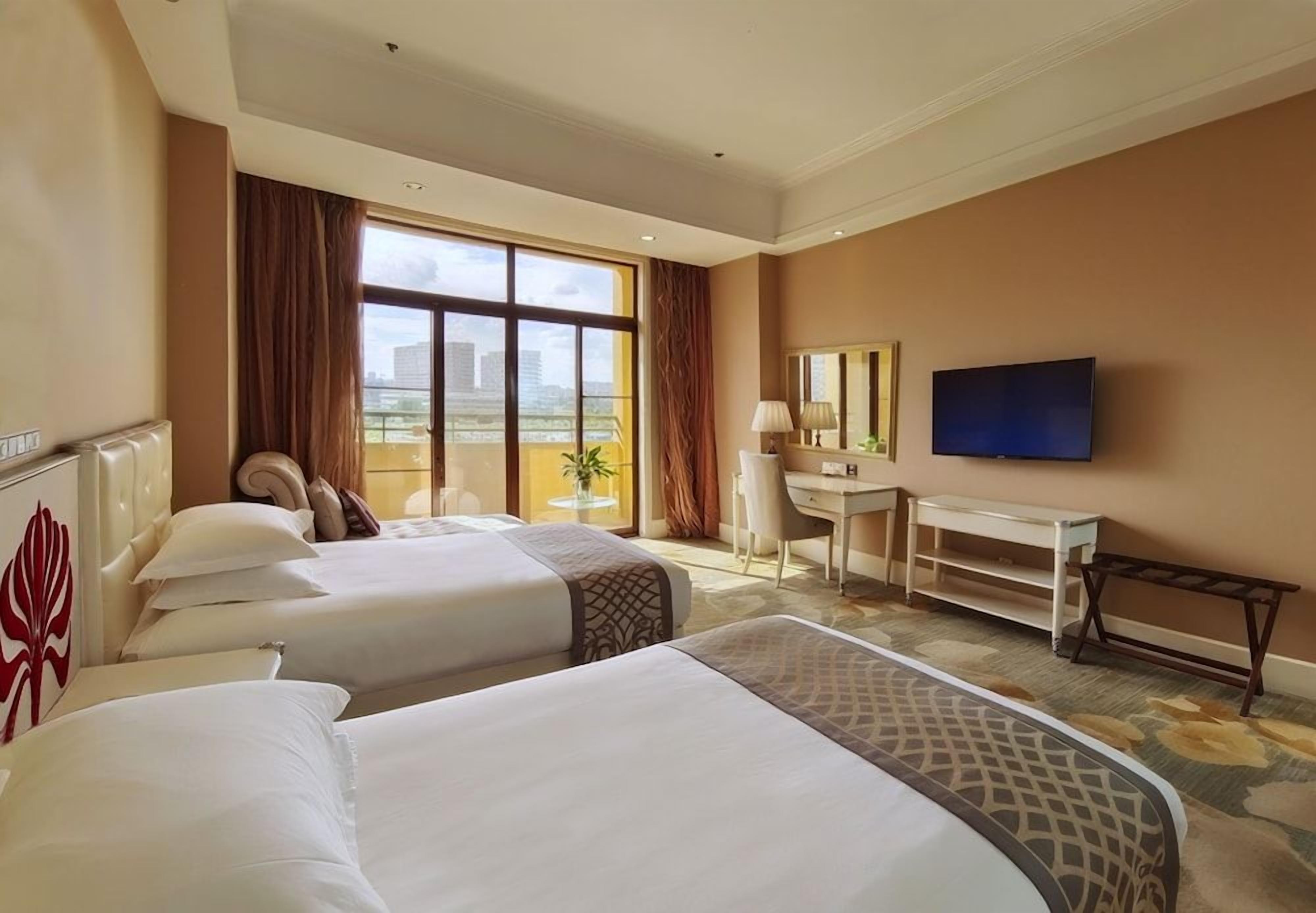 A hotel room with two beds, a desk, a chair, a television, and a large window with curtains.