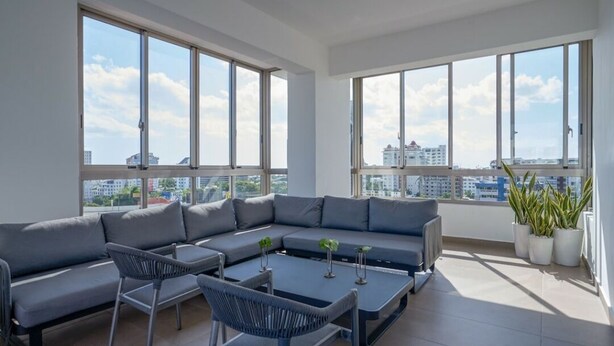 A modern living room with a sectional sofa, two chairs, a coffee table, and large windows offering a city view.