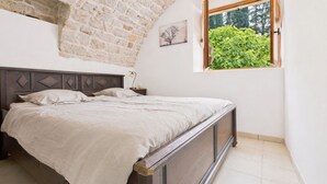 Een slaapkamer met een houten bed, nachtkastje, lamp, raam met uitzicht op groen en een stenen muur.