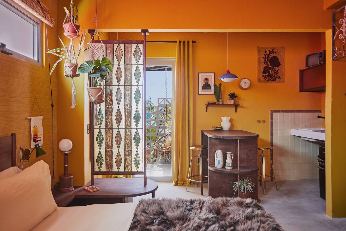 Ein gemütliches Zimmer mit Bett, einem kleinen Tisch, einem Sessel, einer Couch, einem Fellteppich, einer hängenden Pflanze, einem Regal mit Dekoration, einer Wanduhr, einem Fenster mit Vorhängen und Blick auf den Außenbalkon.