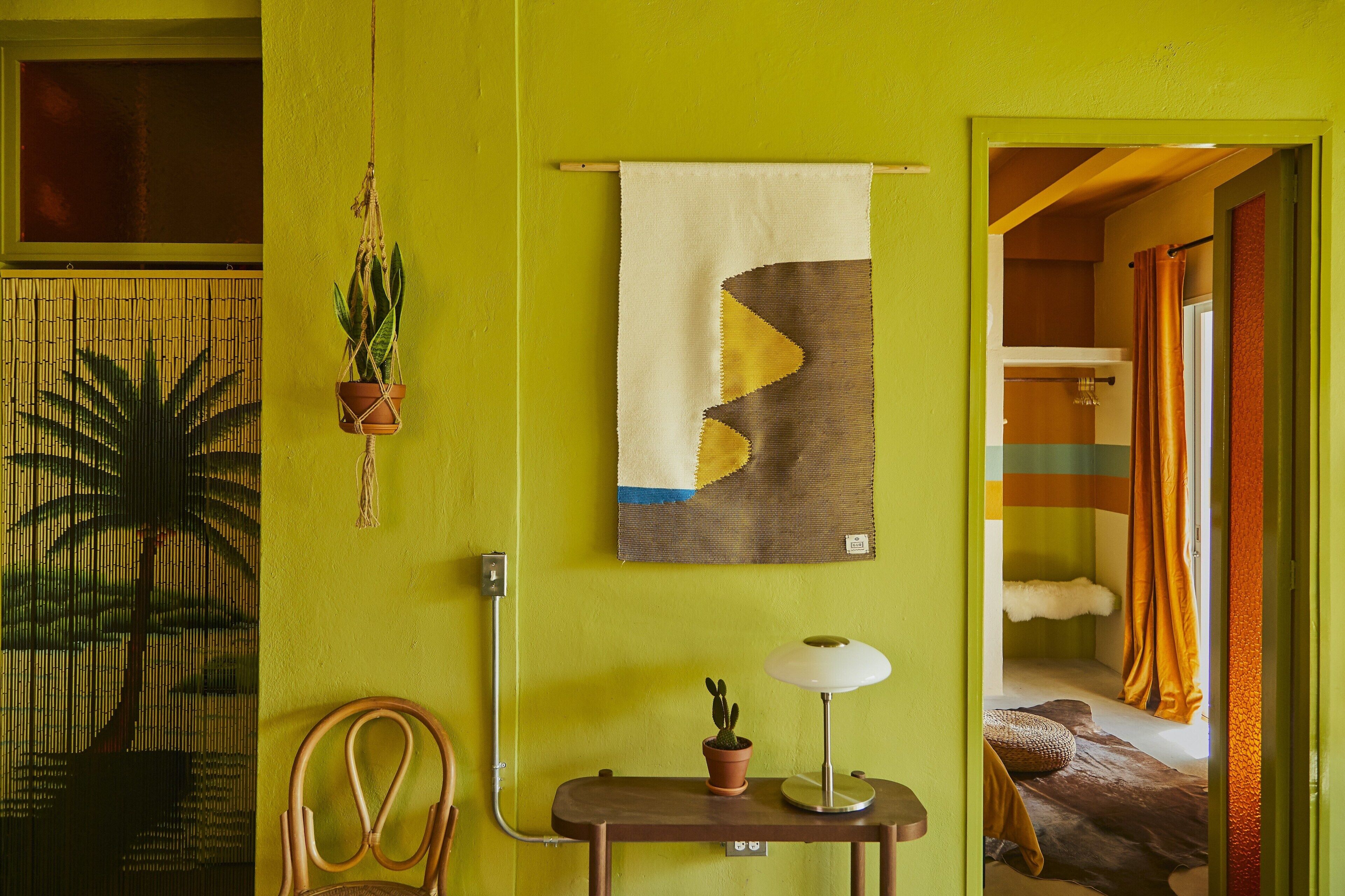 A room with a green wall, a hanging plant, a table with a lamp, and a bed with orange curtains.