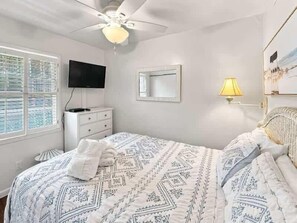 A bedroom with a bed, a television, a dresser, a ceiling fan, a window, and a wall-mounted lamp. - Breakers Villa 221 - Walk to Beach, Shops and Dining (Hilton Head Island)