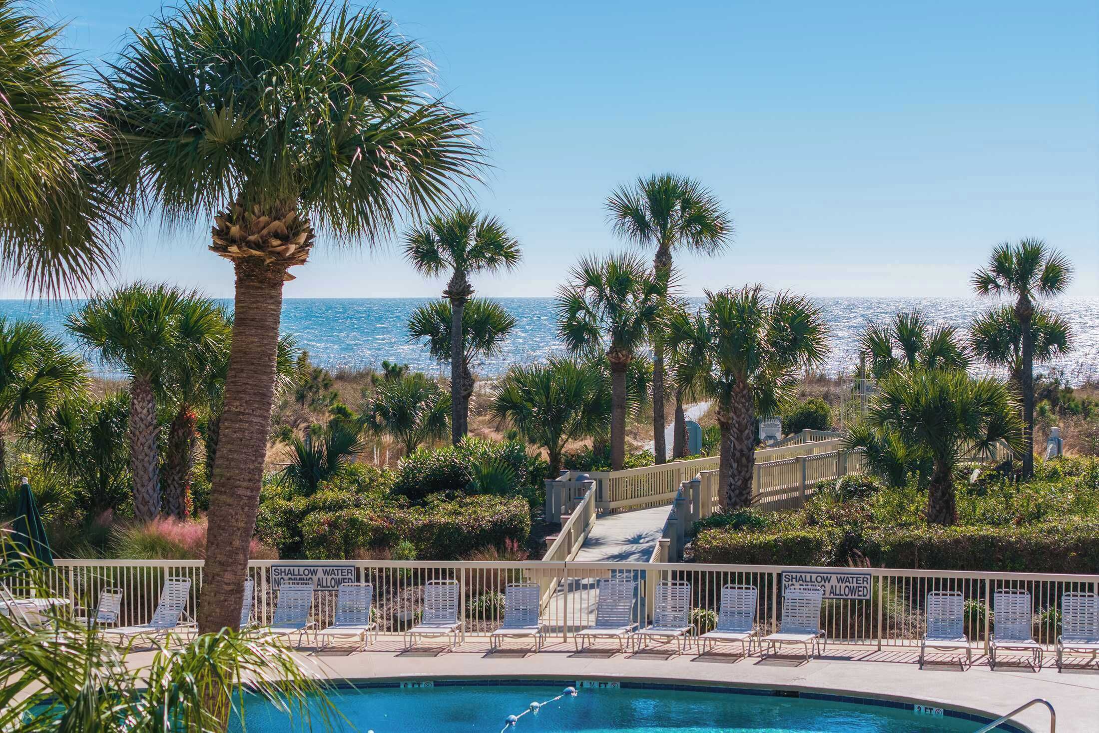 A pool area with lounge chairs, a sign indicating shallow water, and a view of the ocean.
