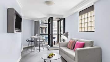 A modern living room with a grey sofa, a coffee table with fruit, a dining area with chairs, and large windows offering a view of the city.