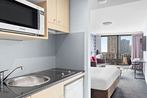A modern kitchen with a microwave, sink, and stove, a bedroom with a bed and a desk, and a view of buildings through large windows. - Mantra Sydney Central (Sydney)