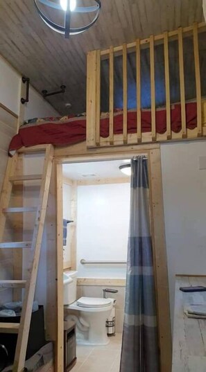 Ett litet rum med en sovplats på ett loft, en trästege, en duschhörna och en toalett.