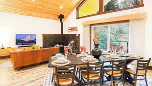 Un coin repas agrémenté d’une cheminée, d’un plafond en bois et d’une grande fenêtre offrant une vue sur les arbres.