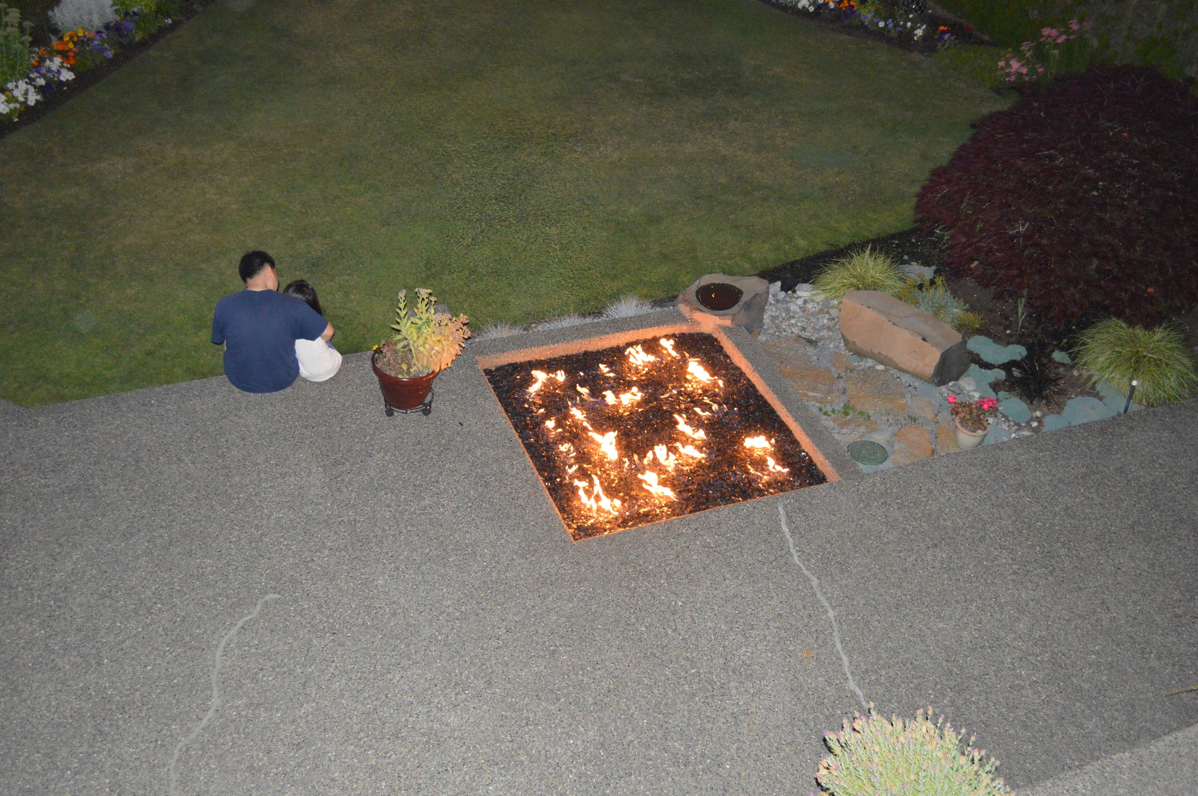 A fire feature in a garden area with a paved surface, potted plants, and a grassy lawn.