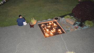 A fire feature in a garden area with a paved surface, potted plants, and a grassy lawn.