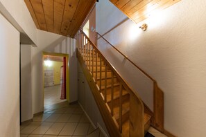 Eine Holztreppe mit Handlauf, die zu einer oberen Etage führt. Die Wände sind weiß gestrichen, und der Boden ist mit Fliesen belegt.