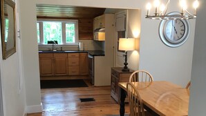 Un plancher en bois mène à un coin repas avec une table et des chaises, une horloge au mur et une cuisine à l’arrière-plan.