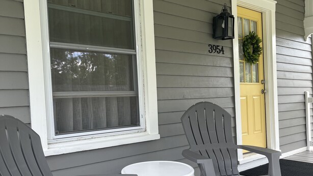 Une véranda avec deux fauteuils Adirondack gris et une petite table blanche. On aperçoit une porte jaune ornée d’une guirlande, et le numéro de la maison, 3954, est affiché sur le mur.