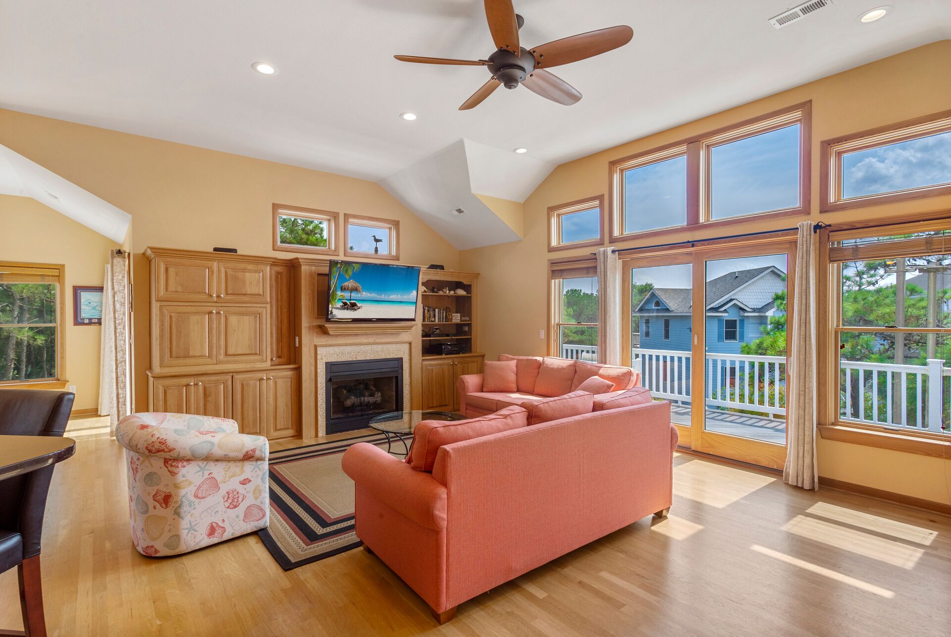 A spacious living room with a fireplace, a large window with a view of a blue house, and a ceiling fan.